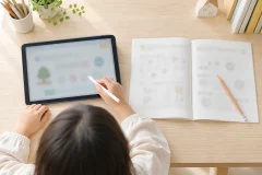 木目の学習机の上に、左に無地のタブレットとタッチペン、右に開いた紙のドリルと鉛筆が並ぶ俯瞰写真
