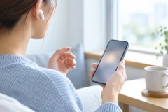 窓辺で片耳にワイヤレスイヤホンをつけ、スマートフォンを手に持つ手元と肩のあたり